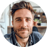 A man with short brown hair and a beard, wearing a denim shirt, smiles at the camera in a well-lit room with shelves and plants in the background.