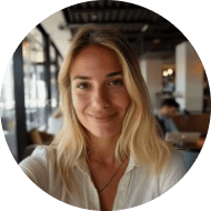 Une femme aux longs cheveux blonds sourit dans un café bien éclairé à l’intérieur moderne. Elle porte une chemise blanche.