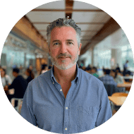 Man with gray hair and beard wearing a blue shirt, standing in a bright, modern indoor space with people seated at tables in the background.