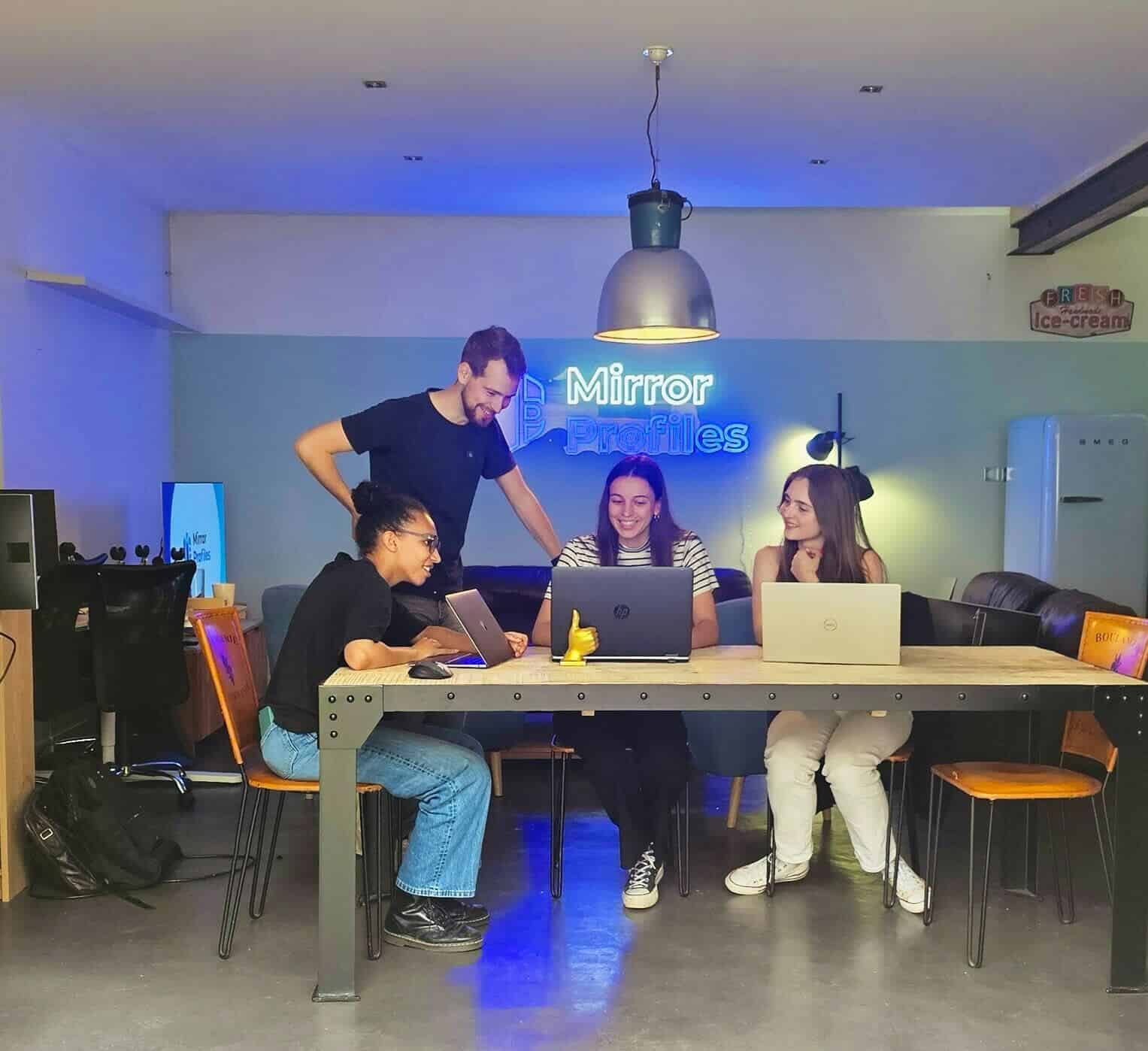 Four people sit and stand around a table with laptops in a modern office space with "Mirror Profiles" signage on the wall.